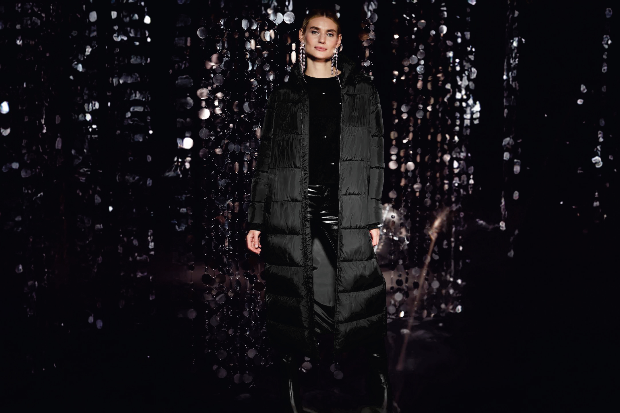 Жена в дълго черно подплатено яке и черни панталони на фона на блестящи пайети.