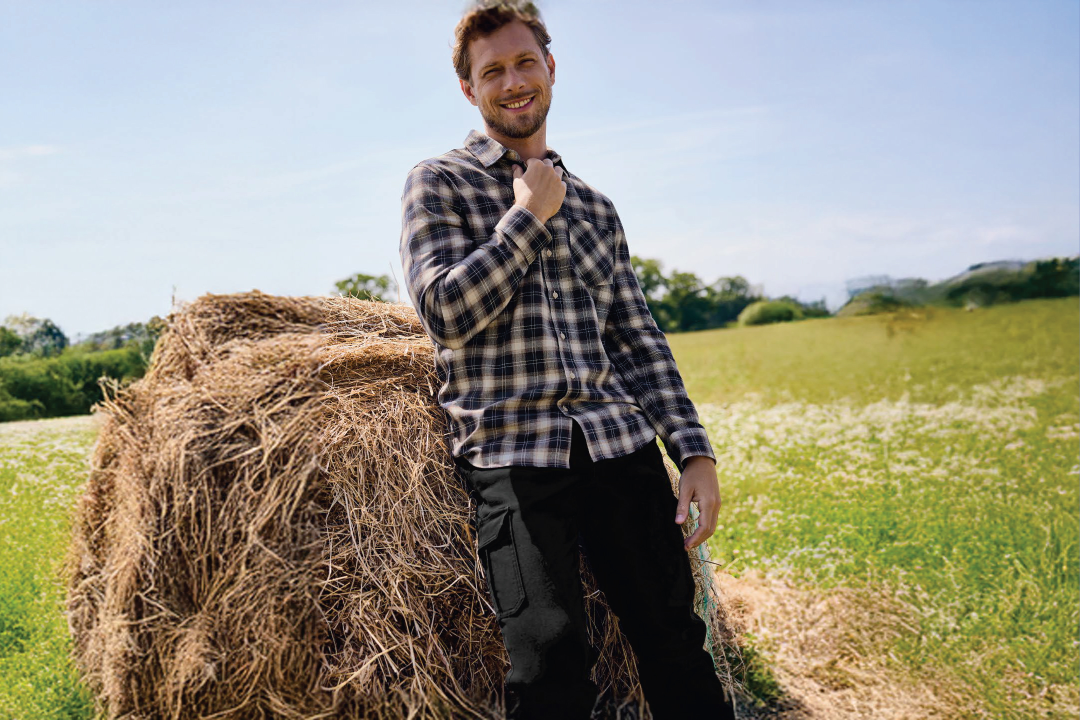 Мъж в карирана риза и черни панталони, облегнат на купа сено в поле.