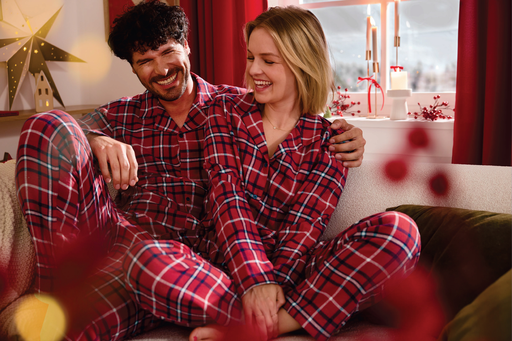 Усмихната двойка в червени карирани пижами, седнала на диван по време на празниците.