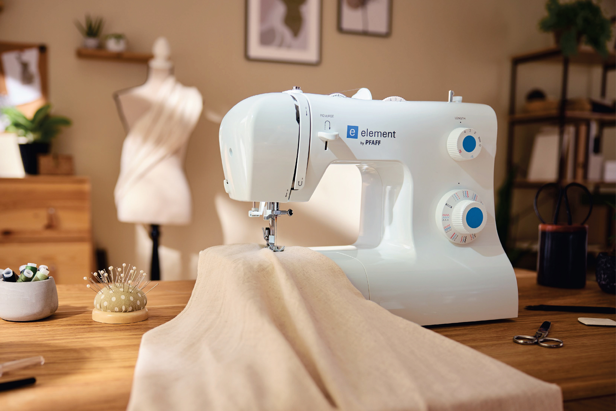 Шевна машина PFAFF Element на дървена маса с шивашки аксесоари и манекен.