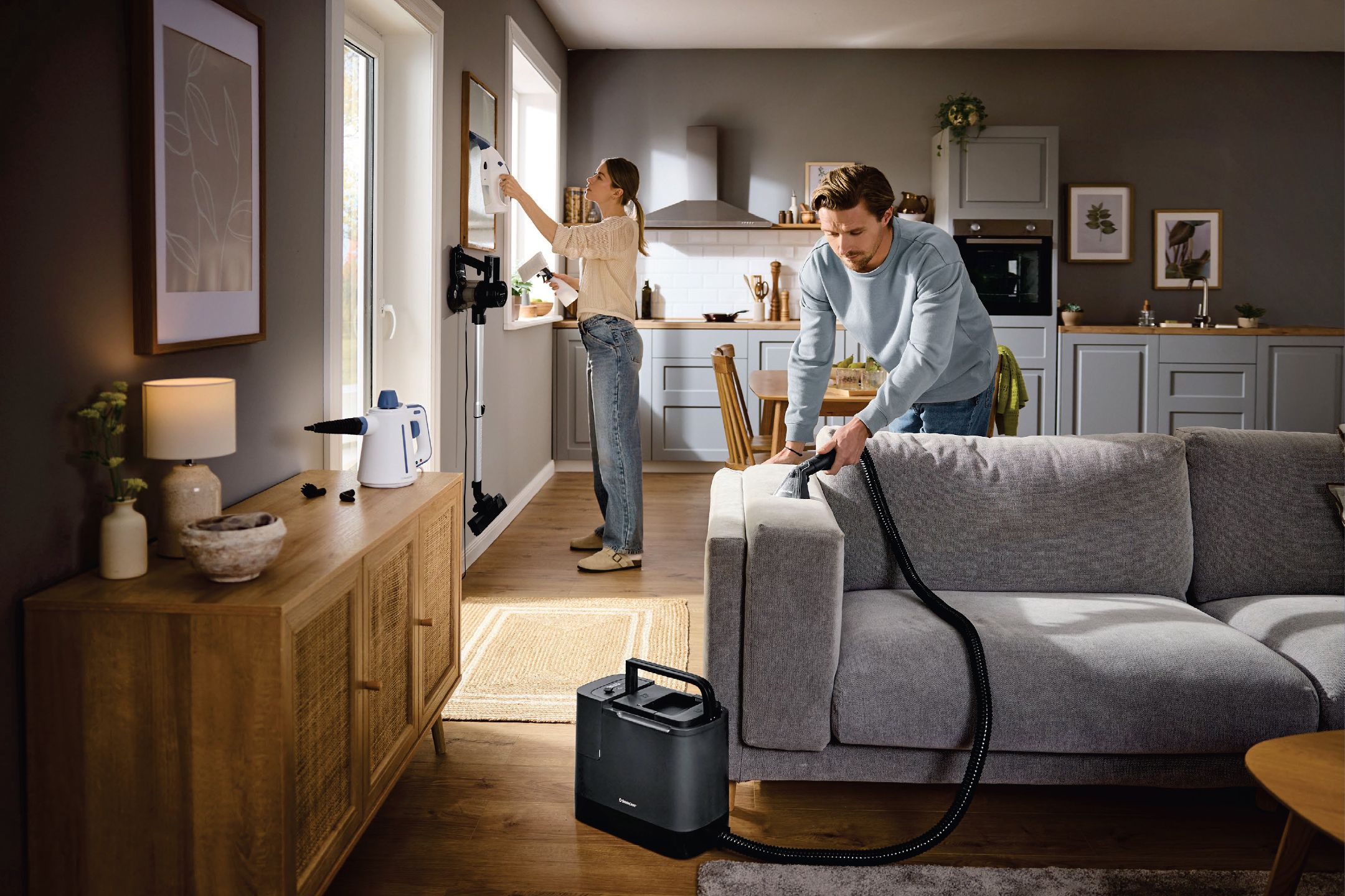 Мъж почиства диван с уред за почистване на тапицерии, докато жена почиства прозорец с парочистачка.