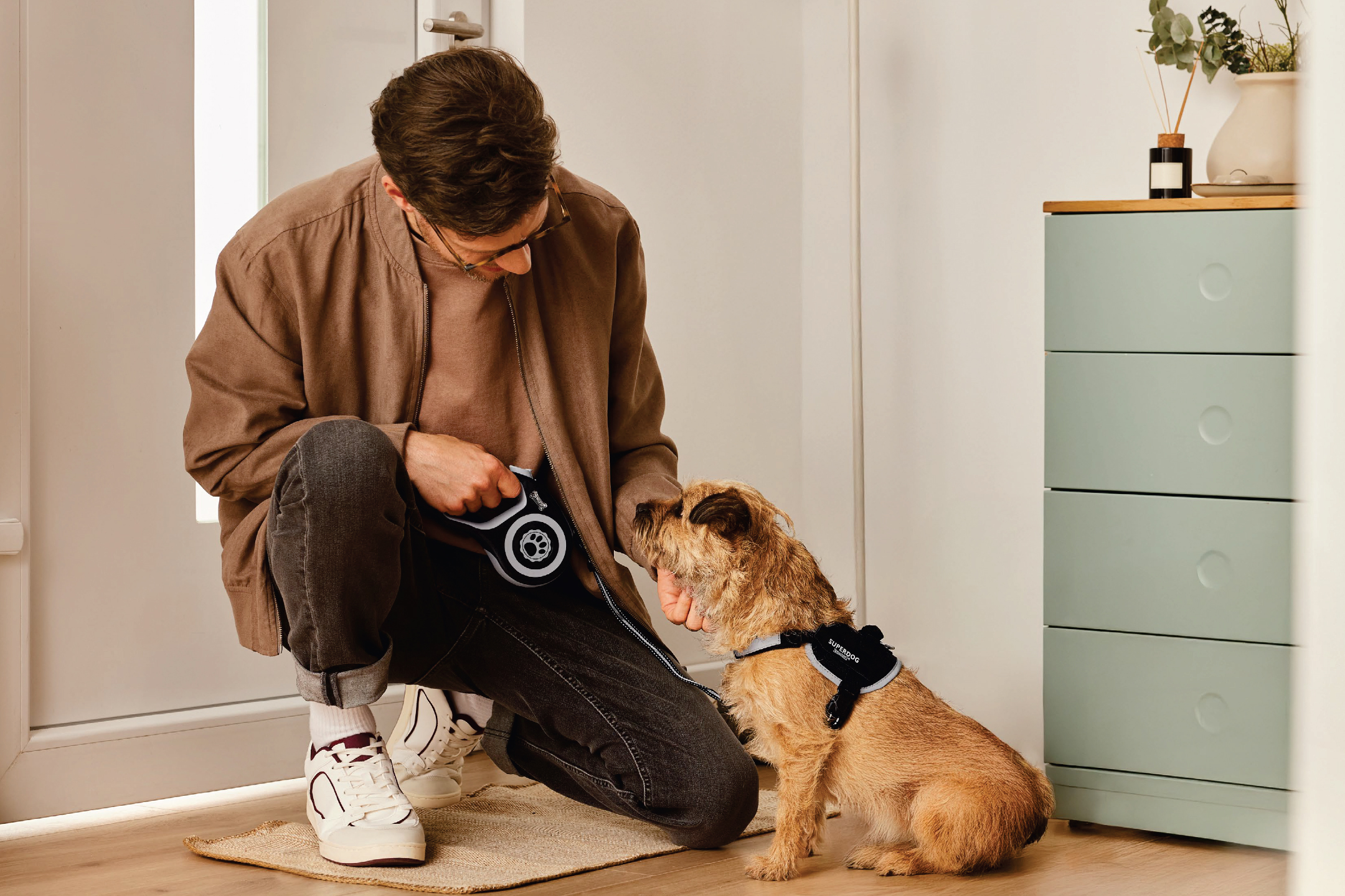 Мъж с кафяво яке и черни дънки, подготвя кучето си за разходка с повод и нагръдник.