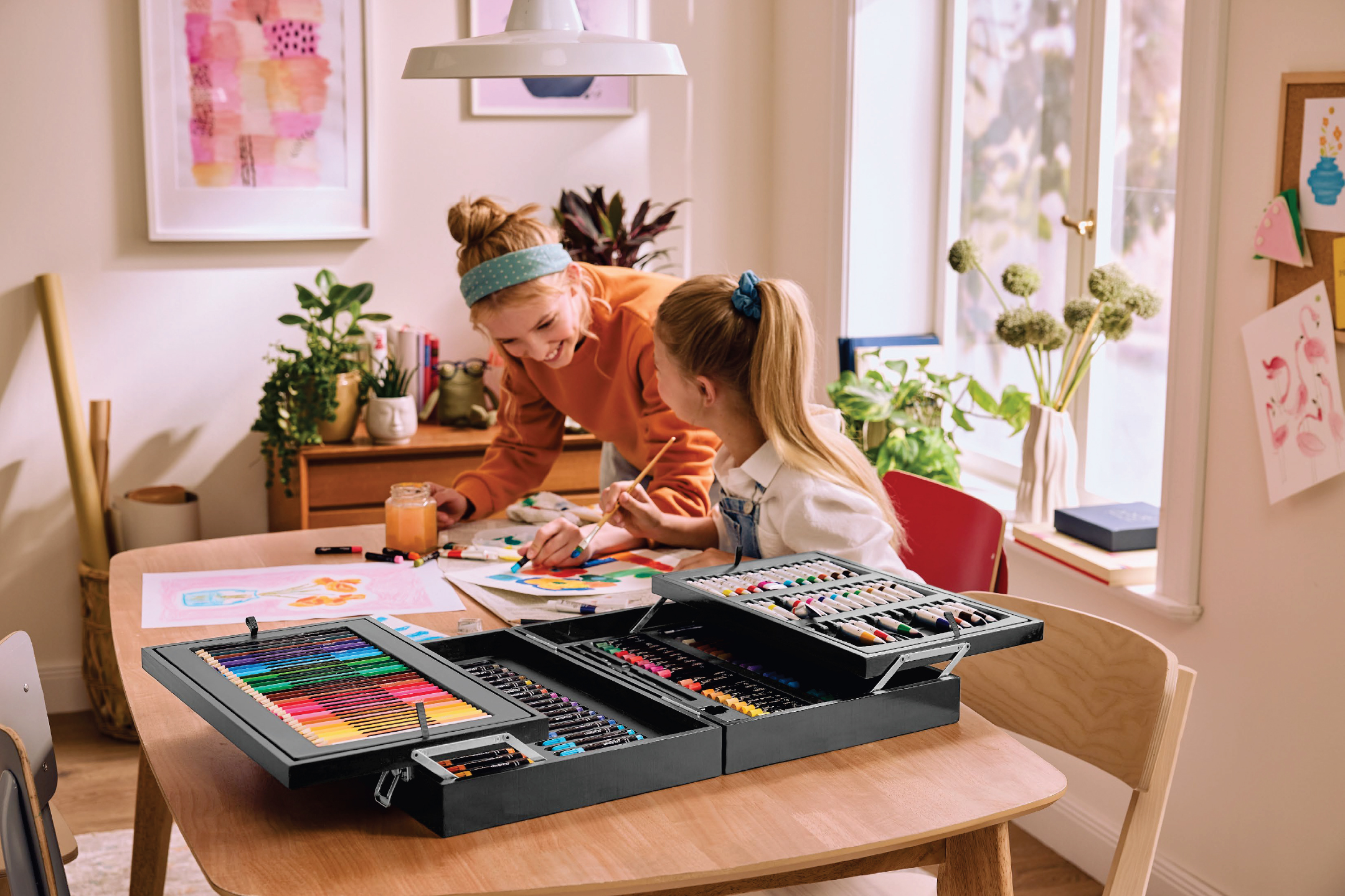 Две момичета рисуват на маса с голям комплект за рисуване, пълен с моливи и бои.