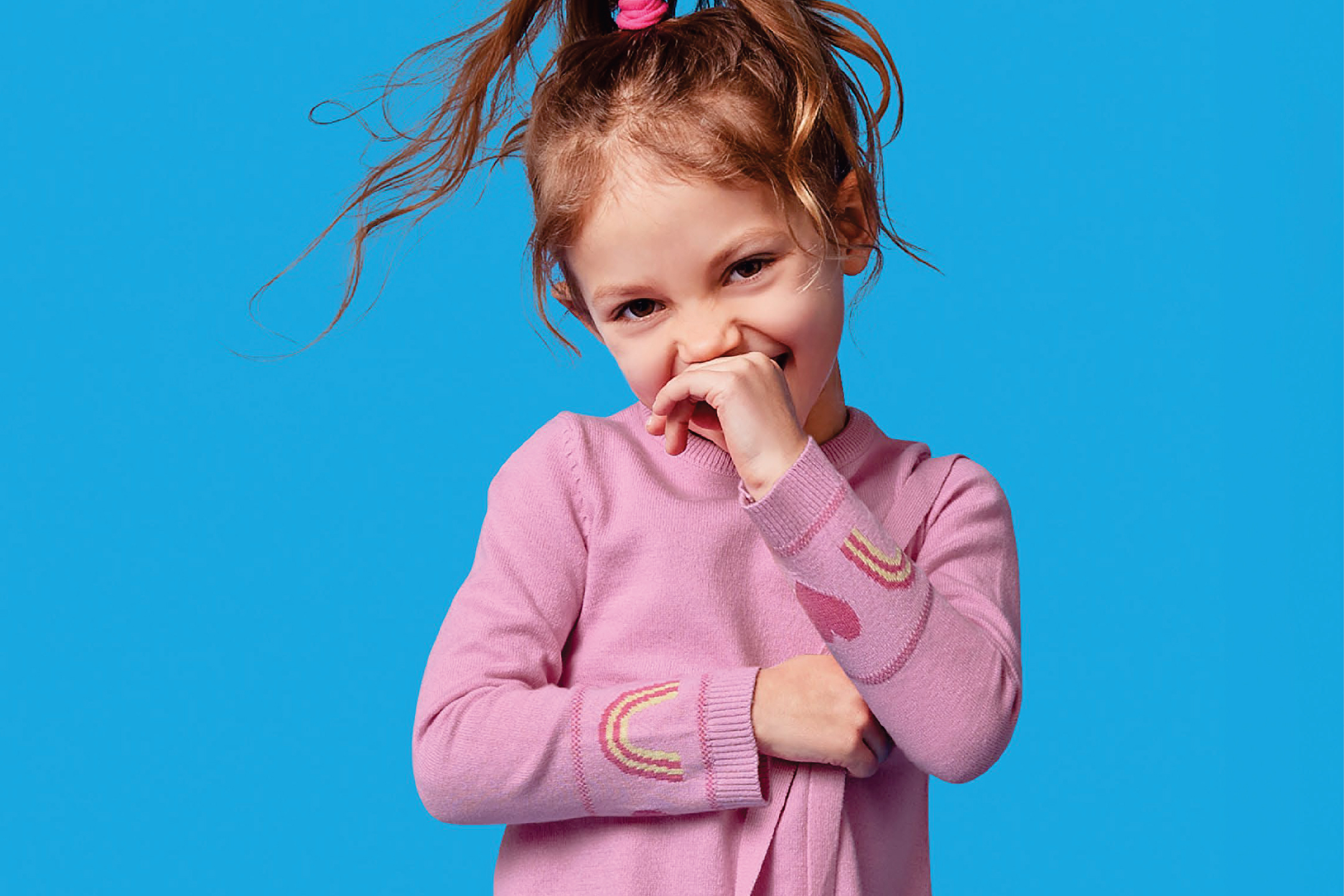 Момиче в розов пуловер с дъги и сърца, прикриващо усмивката си.