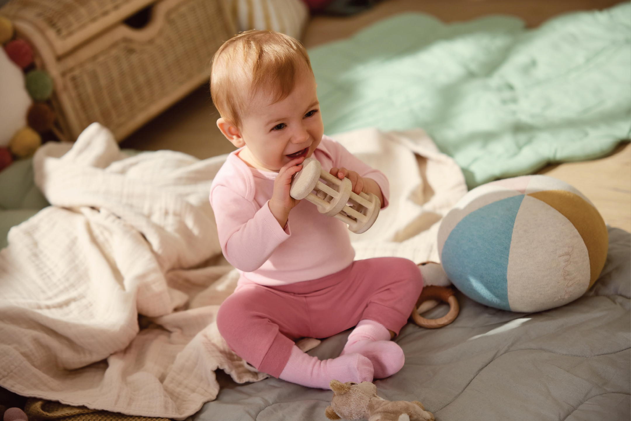Бебе в розови дрехи, седнало на одеяло, играе с дървена дрънкалка.