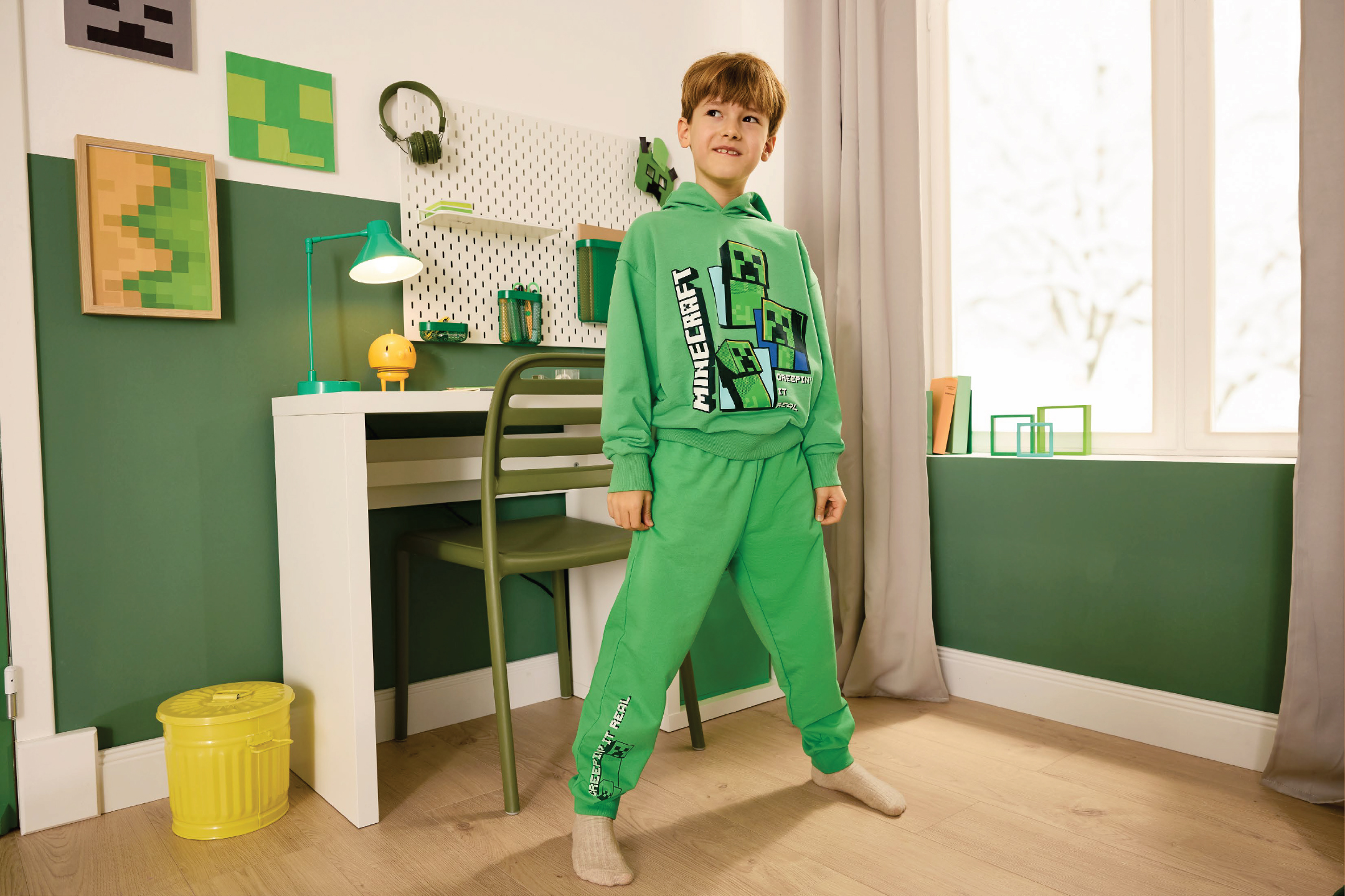 Момче в зелен суичър и панталон с щампа на кубчета, стоящо до бюро в стая с тематична декорация.