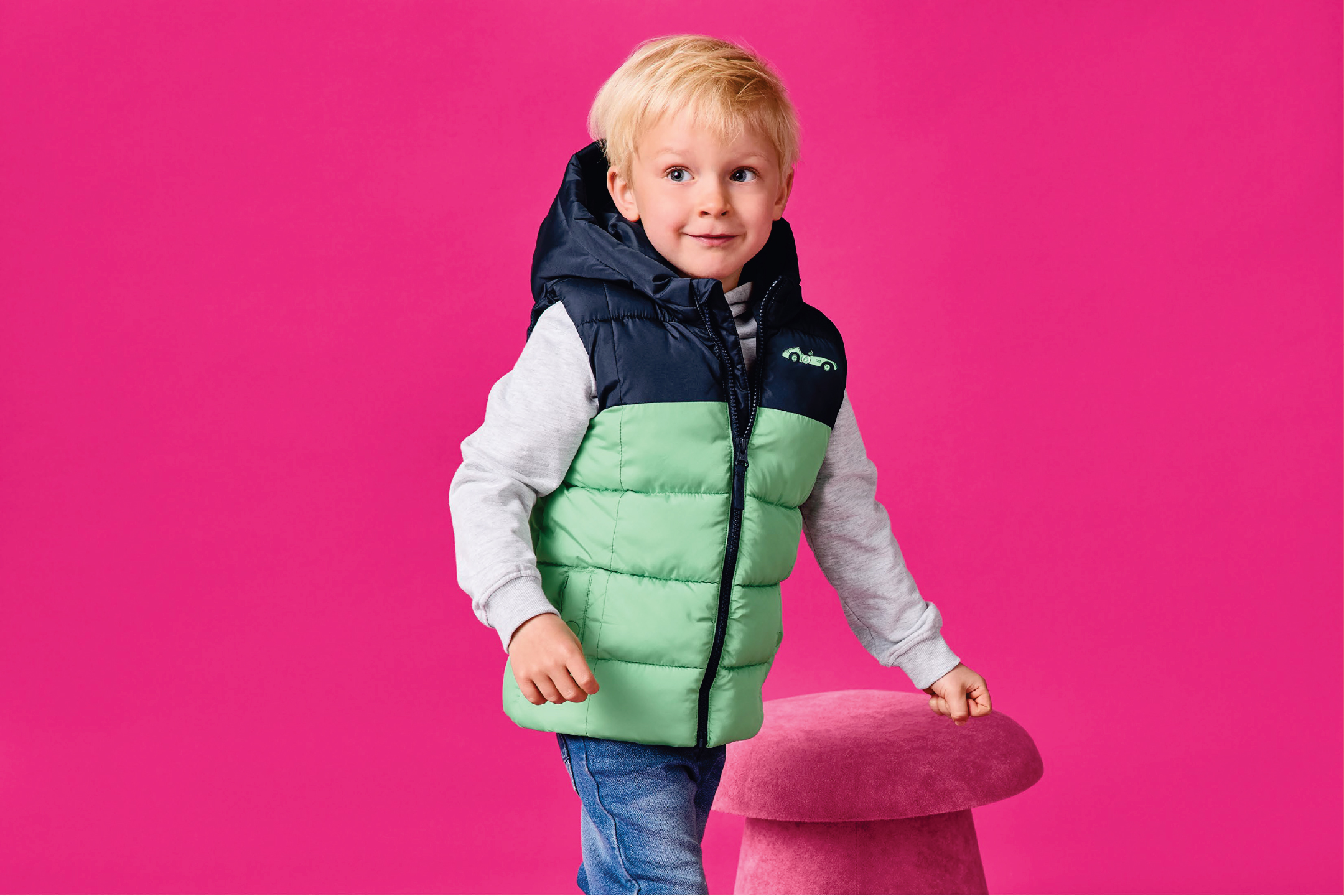 Момче в двуцветен ватиран елек с качулка, сива блуза и дънки, до розова табуретка.