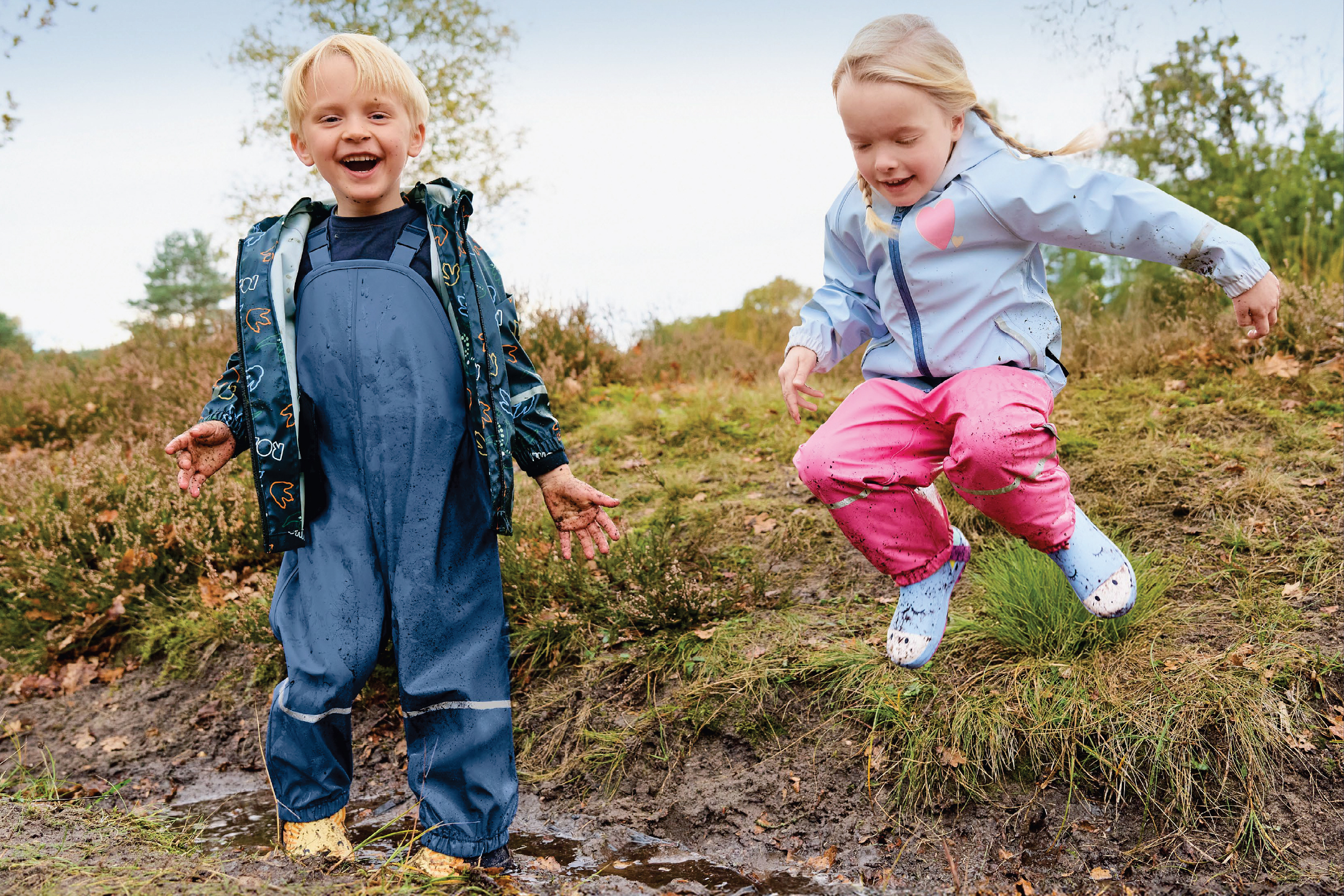 Две деца в непромокаеми гащеризони и якета играят в калта.