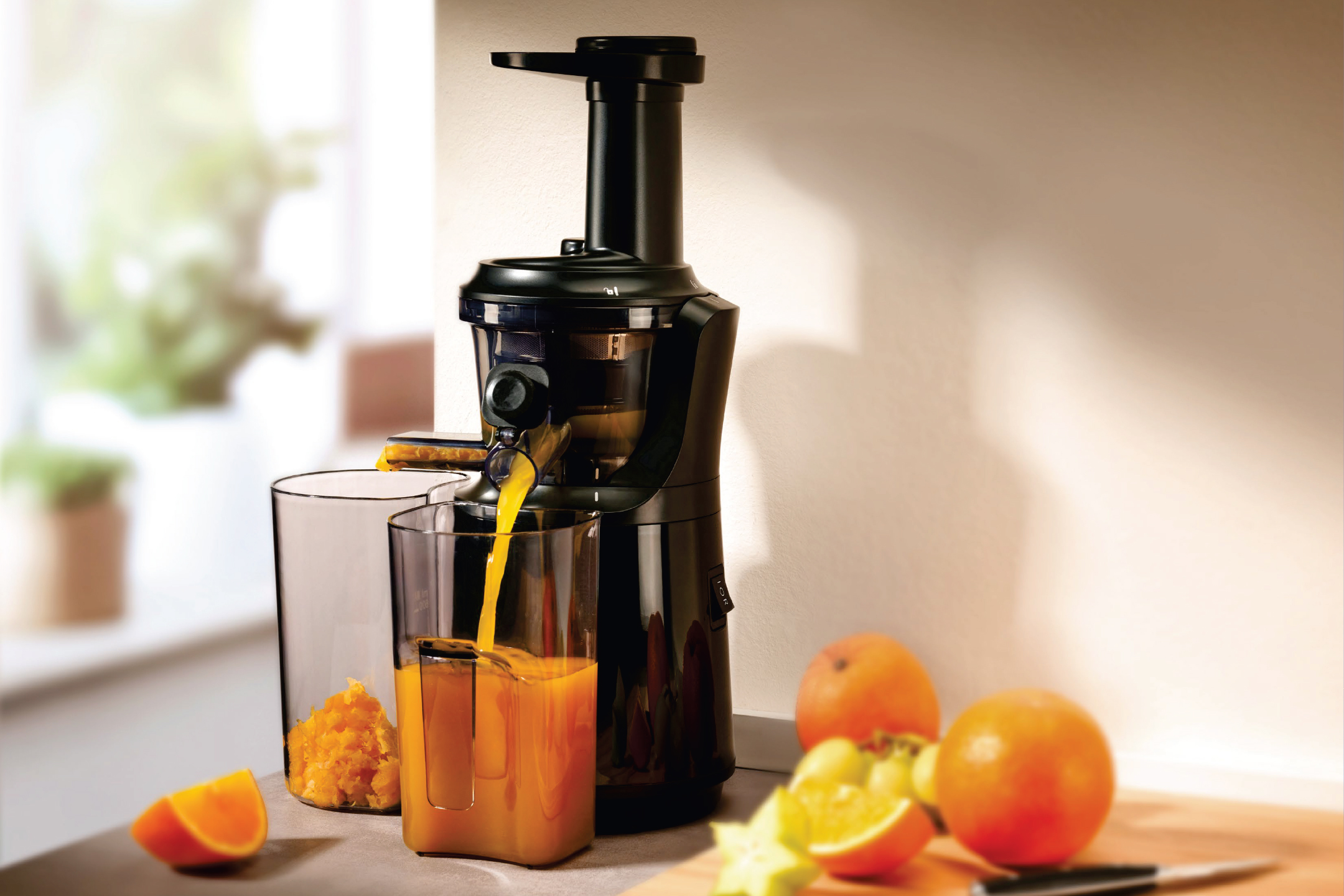 Сокоизстисквачка в действие, пълнеща чаша с портокалов сок, с плодове наблизо.