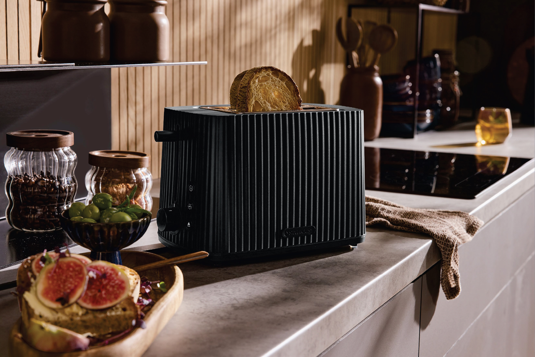 Черен тостер с филия хляб, заобиколен от бурканчета с кафе, маслини и смокини.