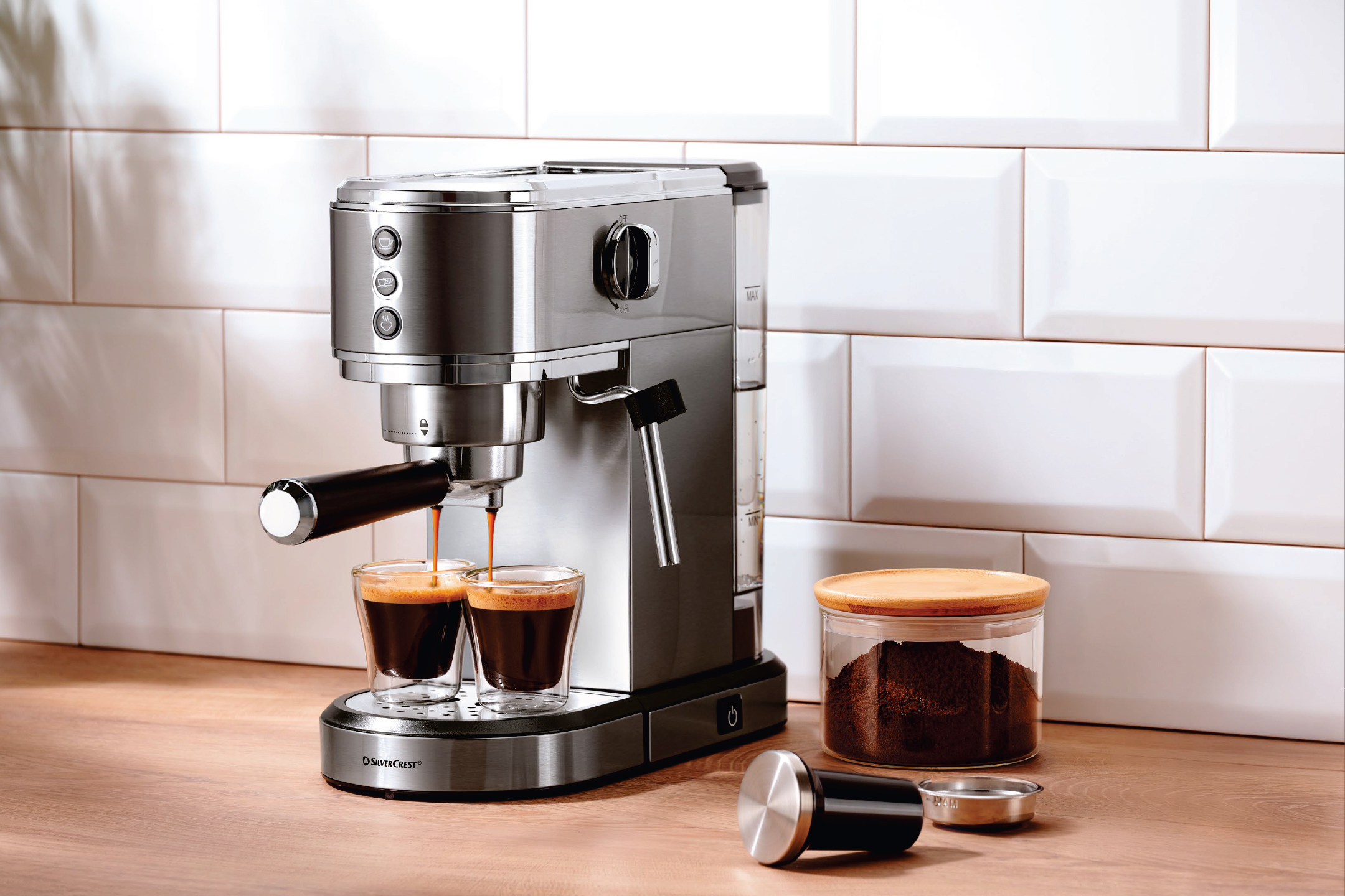 Еспресо машина, пълнеща две чаши кафе, с бурканче смляно кафе на дървен плот.