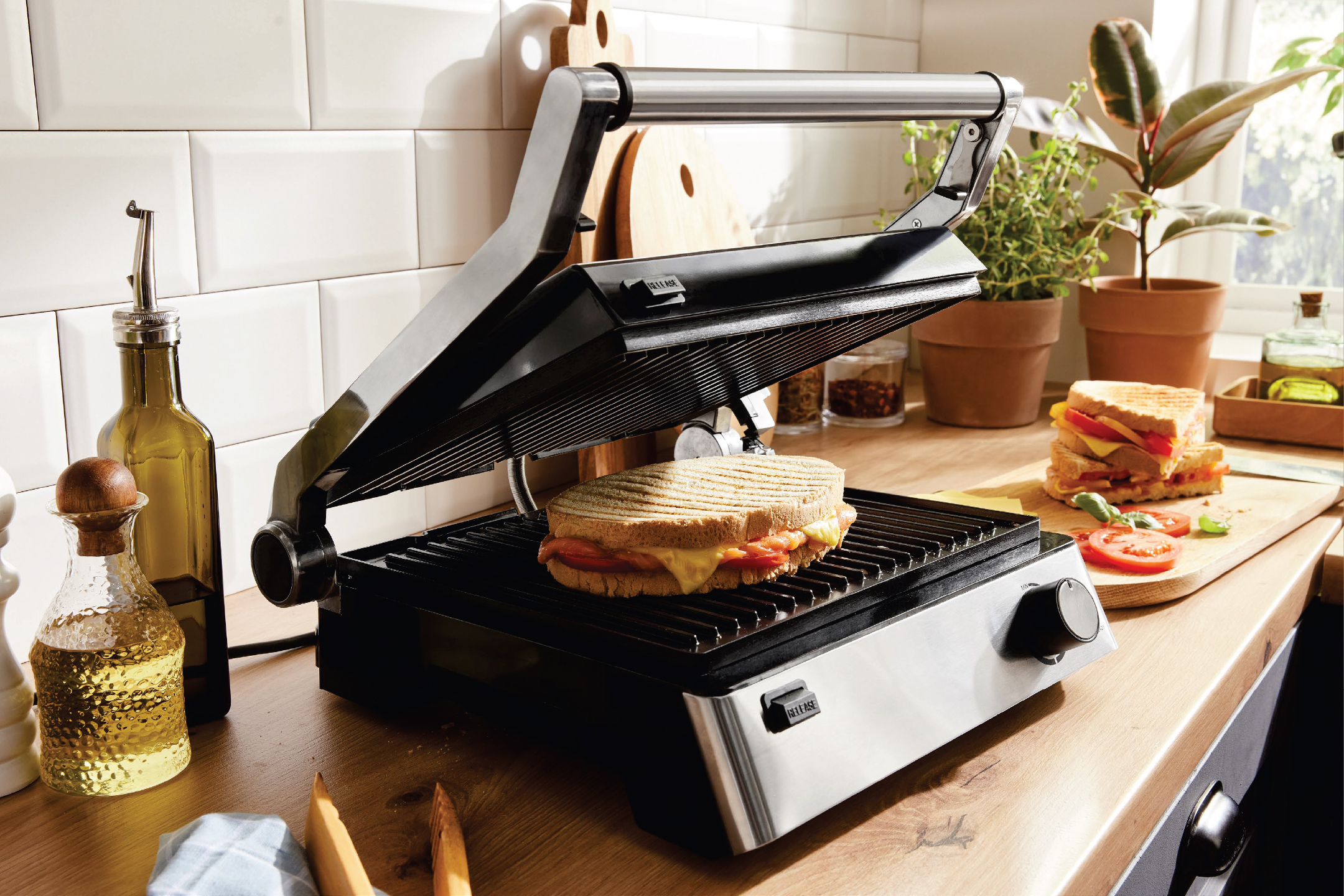 Грил преса с топъл сандвич, бутилки с олио и подправки на кухненски плот.