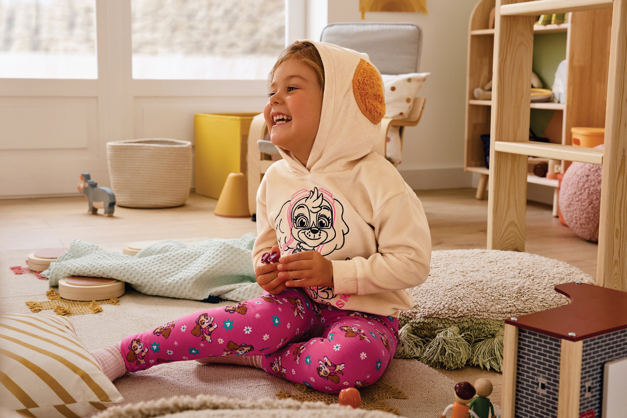 Момиченце в суичър и клин с кученца от Paw Patrol, седнало на килим в детска стая.