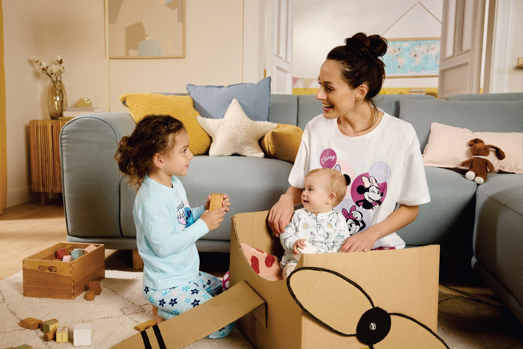 Майка и две деца си играят с картонена кутия, превърната в самолет, и дървени блокчета.