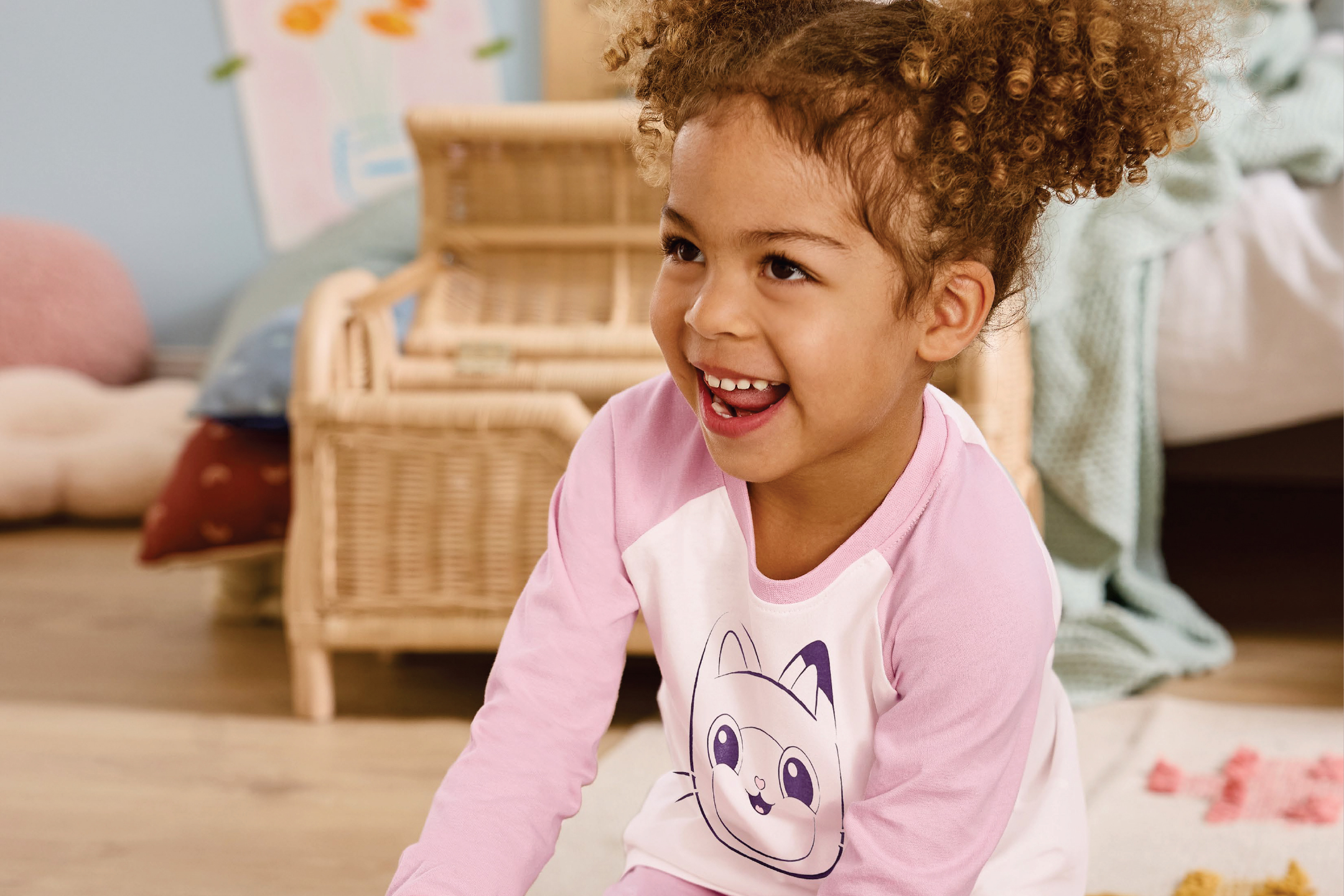 Усмихнато момиченце с къдрава коса, облечено в розова пижама с котешка щампа.
