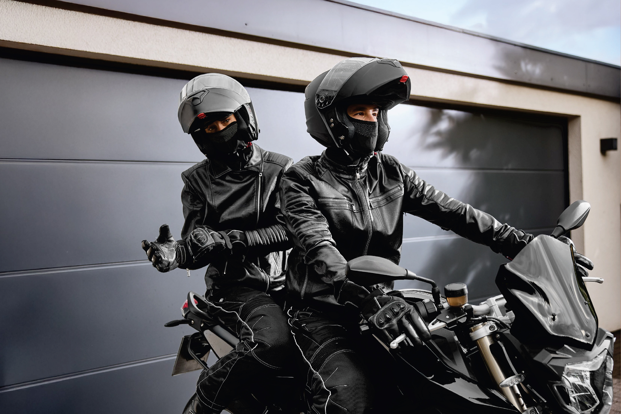 Двама мотористи с кожени якета, каски и балаклави на мотор пред гараж.