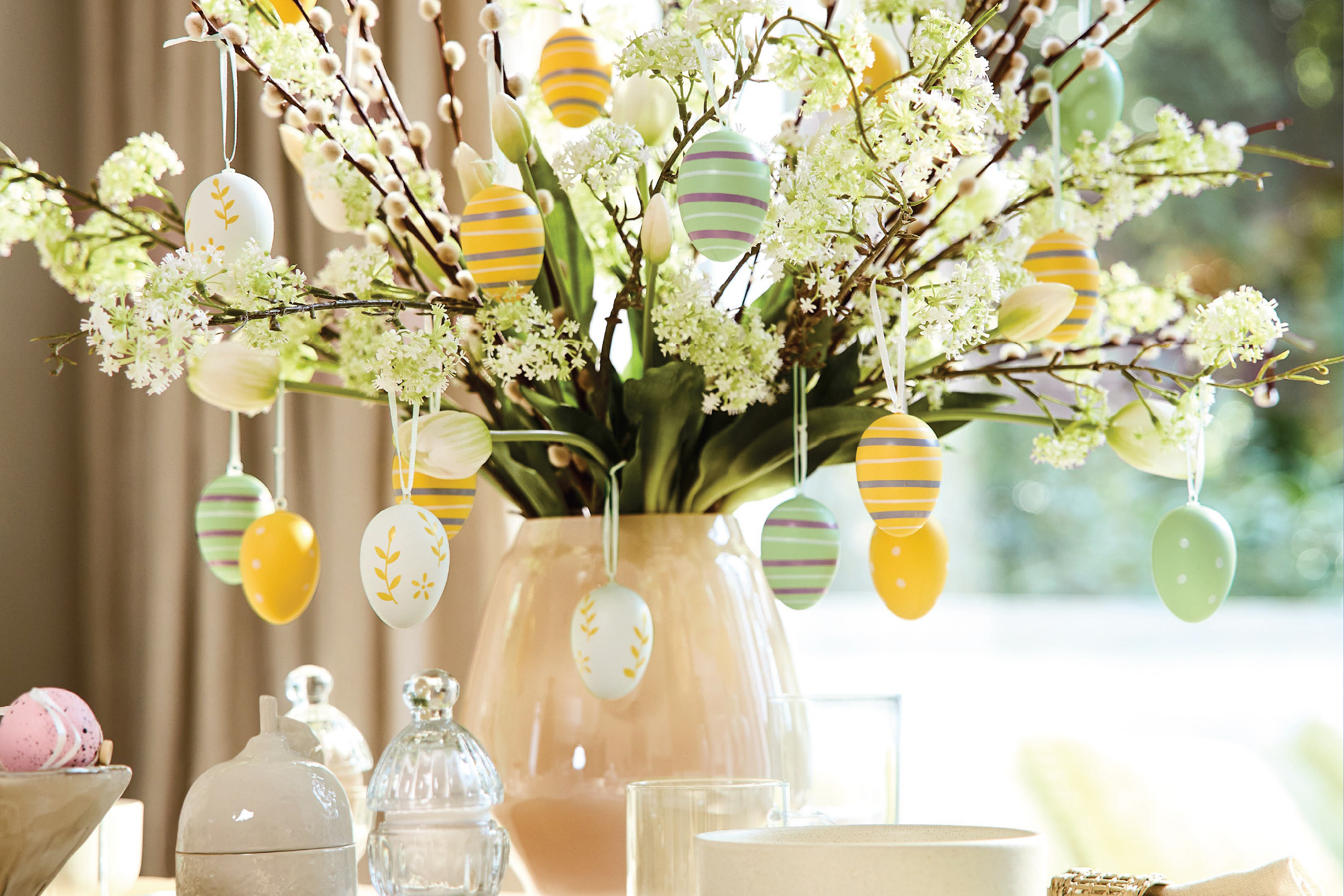 Великденска украса с пъстри яйца, висящи от клонки върба и цветя във ваза.