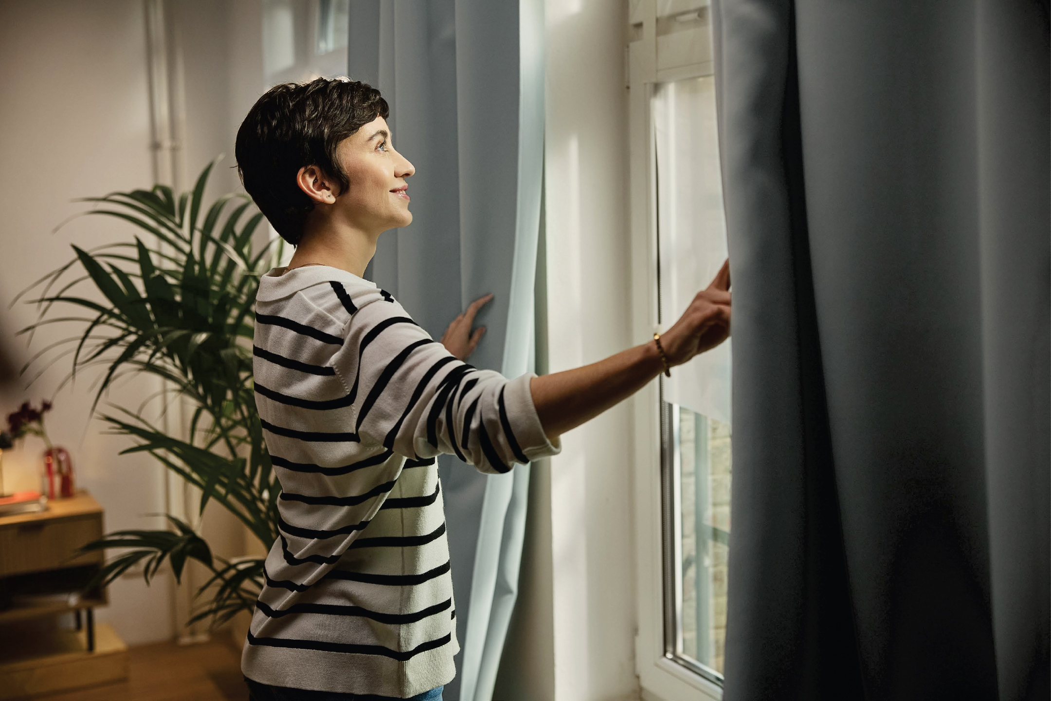 Жена отваря завеси, за да пусне светлина в стая с палма.