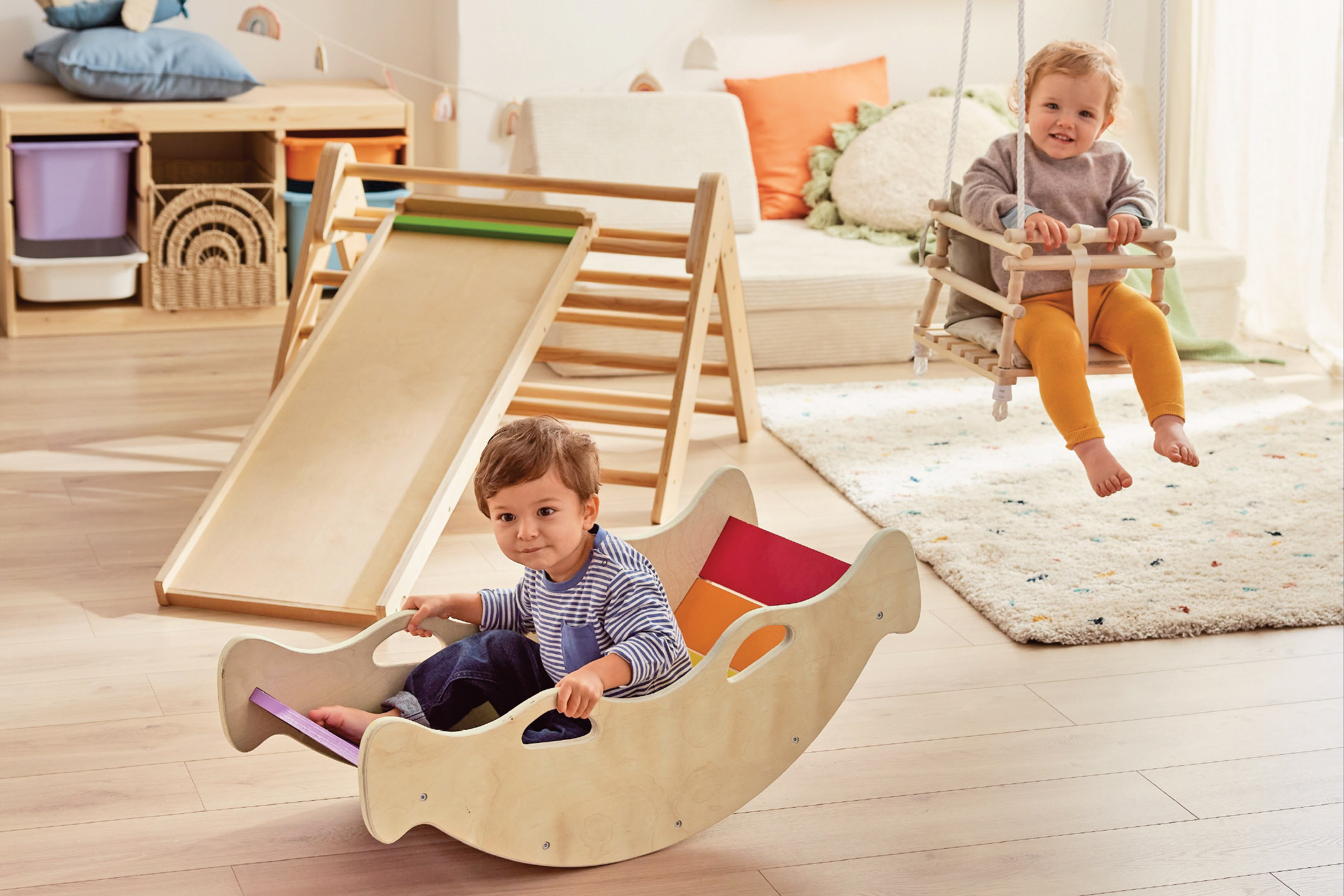 Две бебета си играят с дървена люлка и дървена пързалка в детска стая.