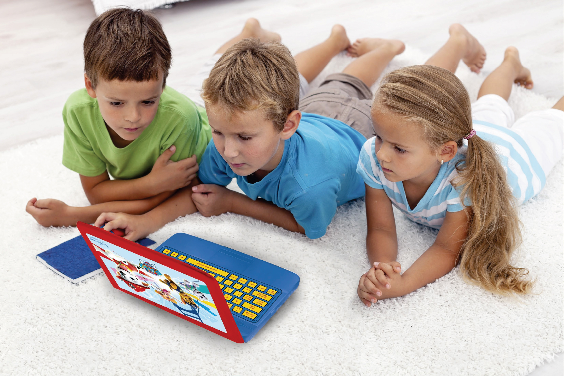 Три деца лежат на килим, гледайки детски лаптоп с анимационни герои.