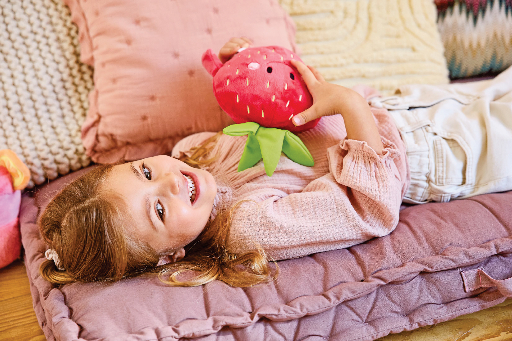 Усмихнато момиченце лежи на розова възглавница, прегърнало плюшена ягода.