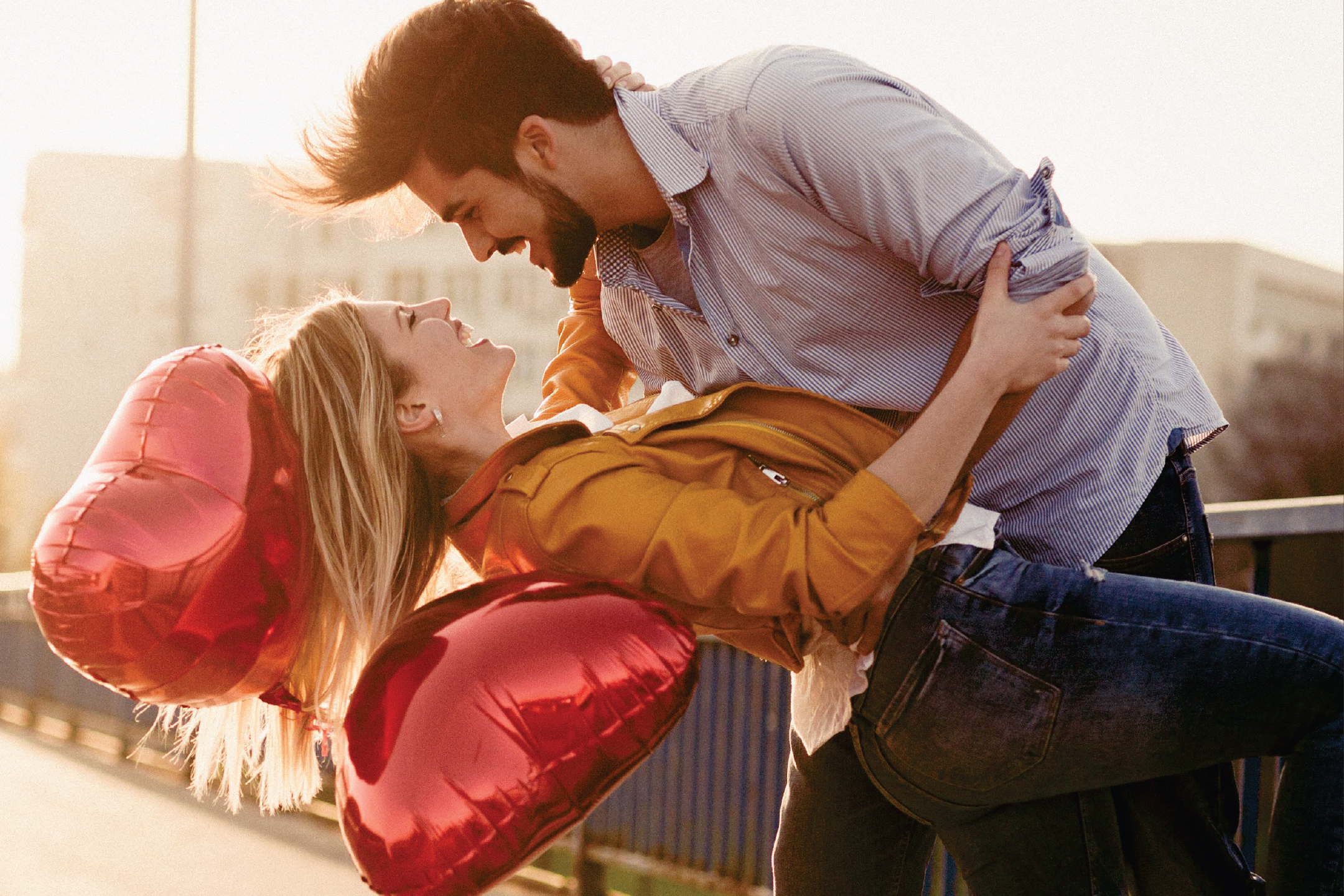 Влюбена двойка се смее, докато мъжът държи жената, заобиколени от червени балони във формата на сърце.