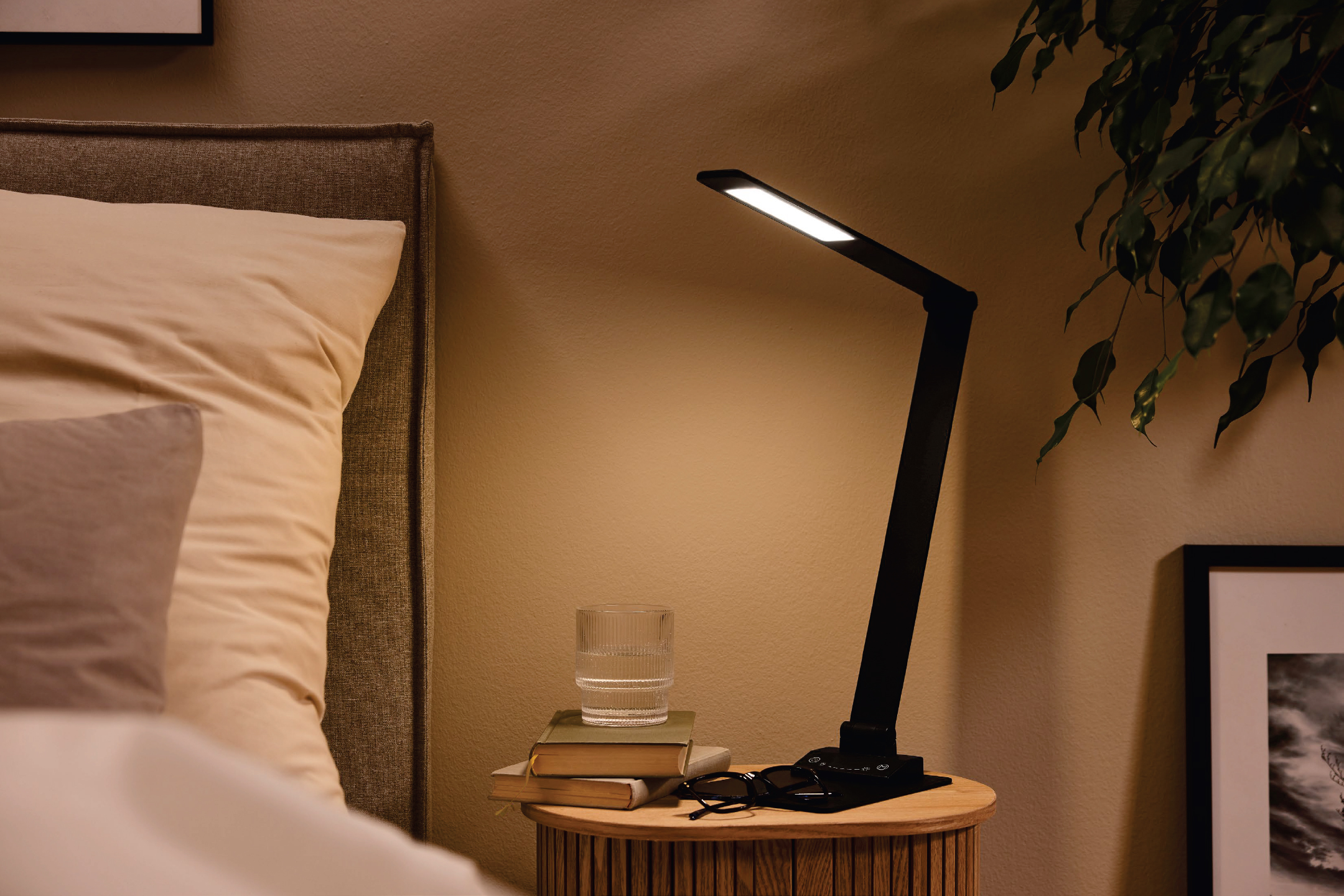 Черна настолна лампа на нощно шкафче до легло с възглавници и стайно растение.