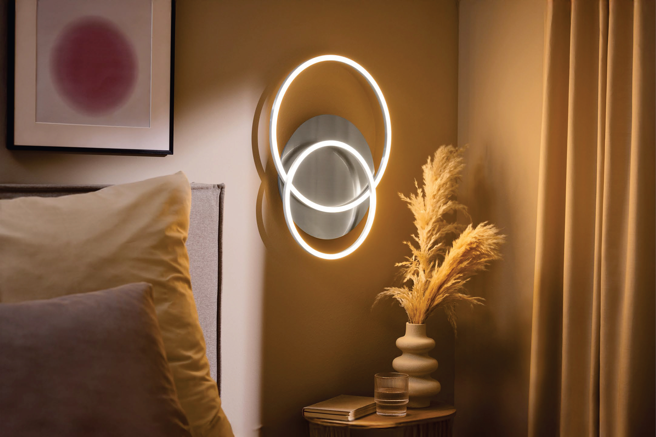 Модерна стенна лампа с кръгове, ваза със сухи цветя и книга на нощно шкафче до легло.