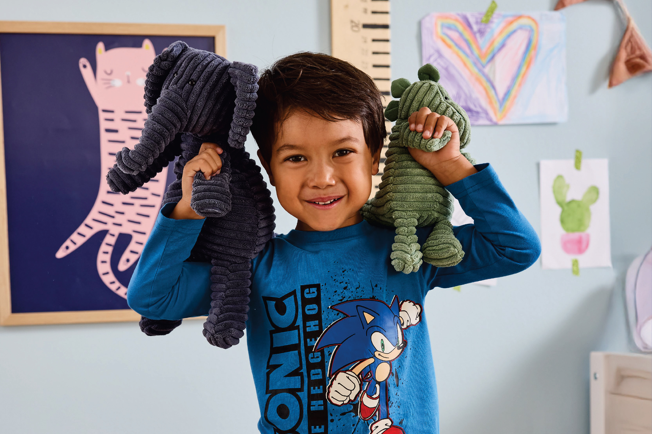 Момче в тениска със Соник, държащо плюшени играчки слон и динозавър.