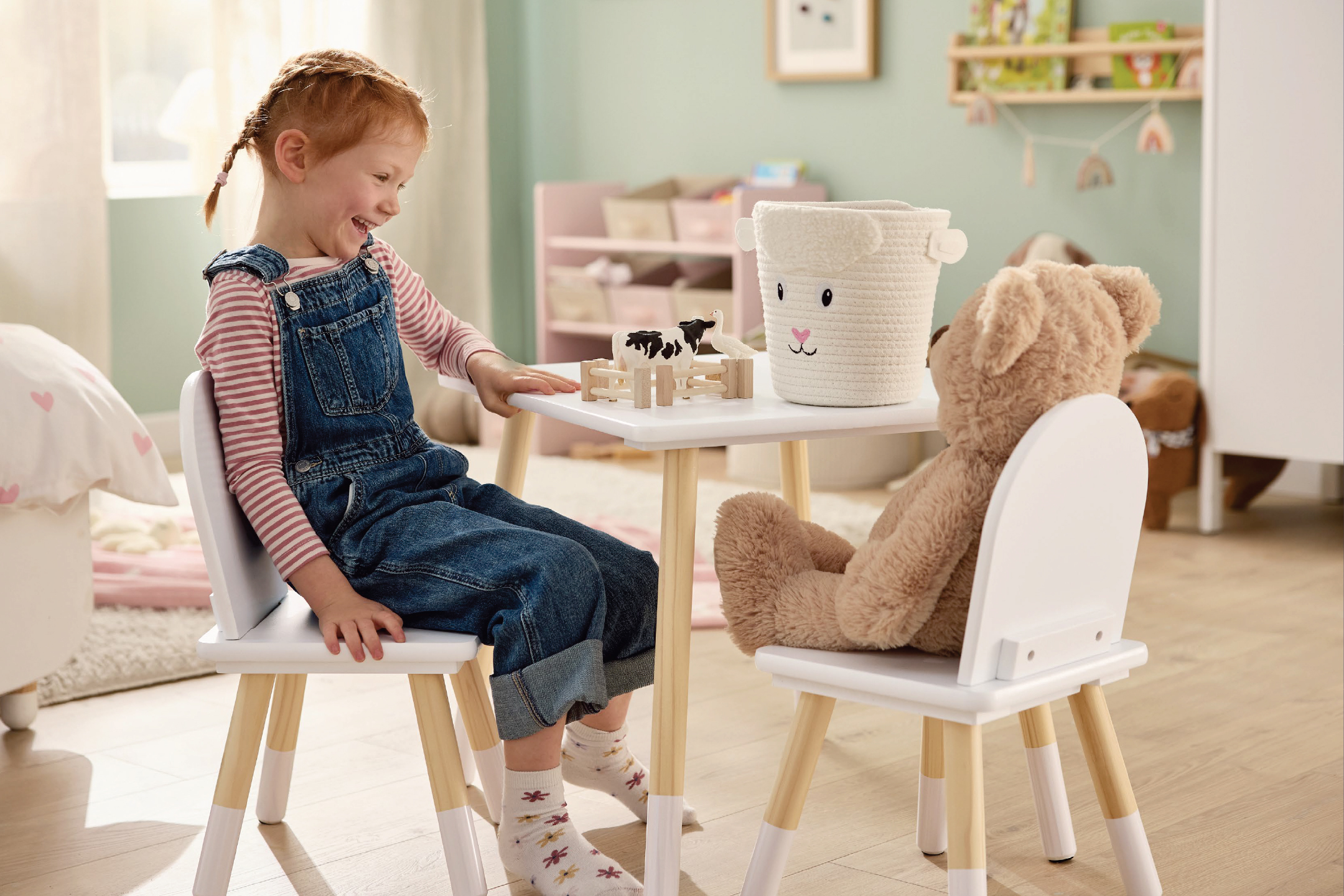 Момиченце и плюшено мече седят на бяла маса и столове в детска стая.