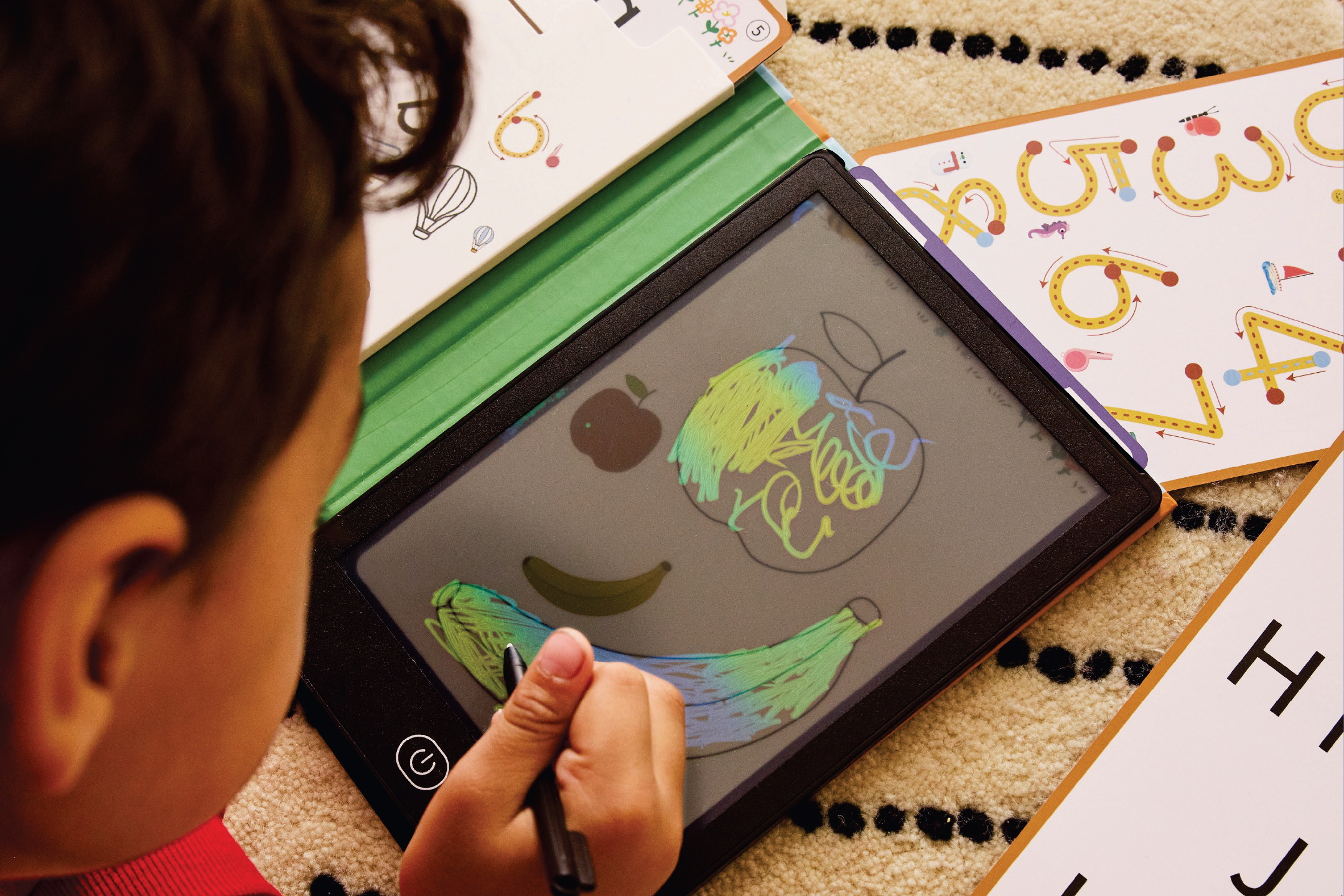 Дете рисува плодове на дигитална дъска, заобиколена от образователни карти.