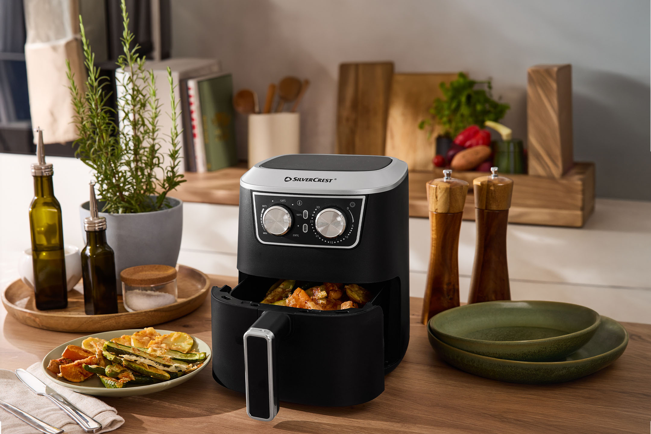 Фритюрник с горещ въздух с печени зеленчуци, зехтин, сол и черен пипер на кухненски плот.