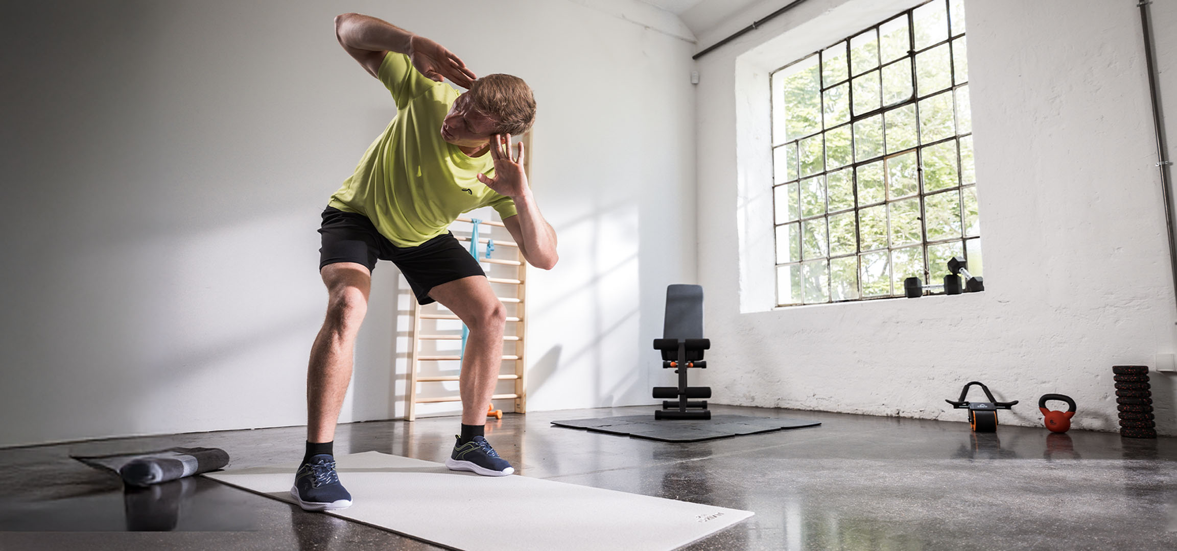 Мъж тренира във фитнес зала, използвайки постелка за йога и дъмбели.