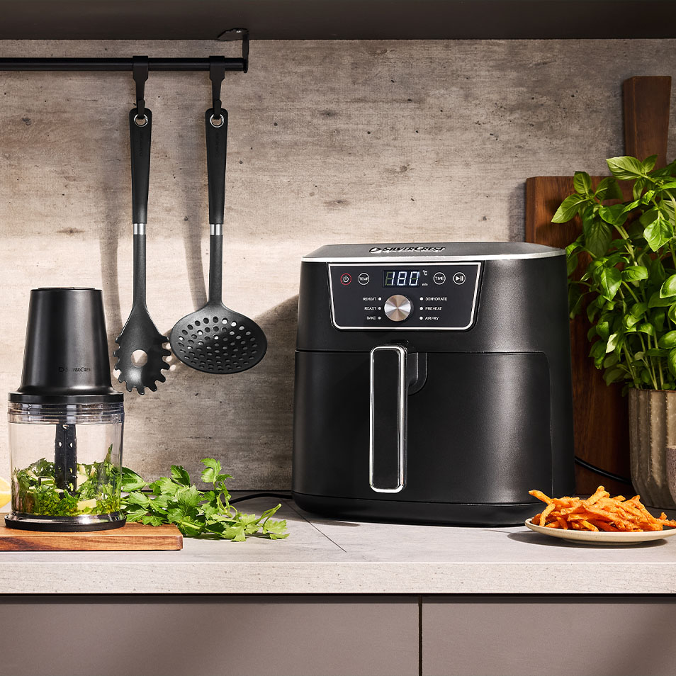 Фритюрник с горещ въздух SilverCrest и чопър за храна на кухненски плот със свежи билки и пържени сладки картофи.