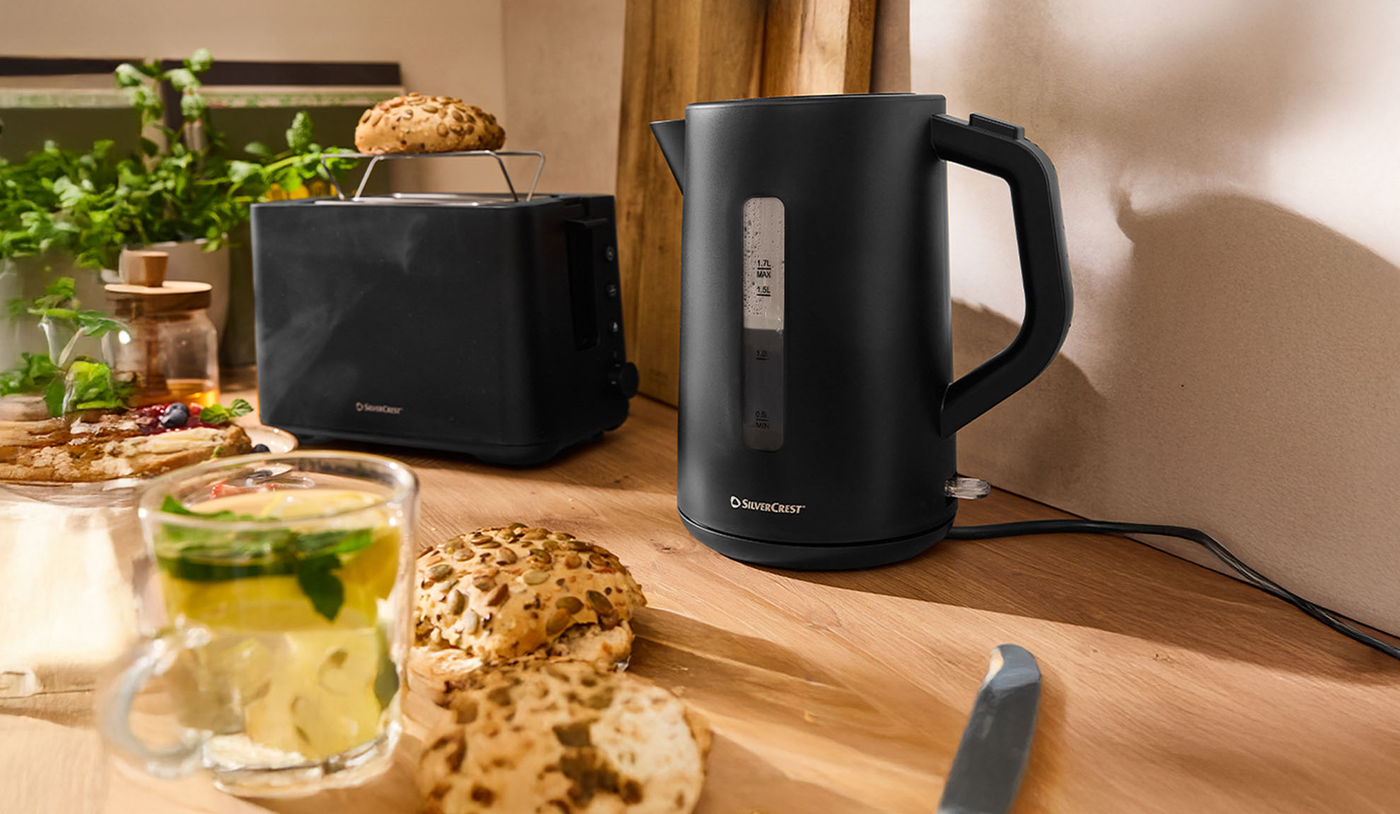 Черен SilverCrest чайник и тостер на дървена маса със закуска.