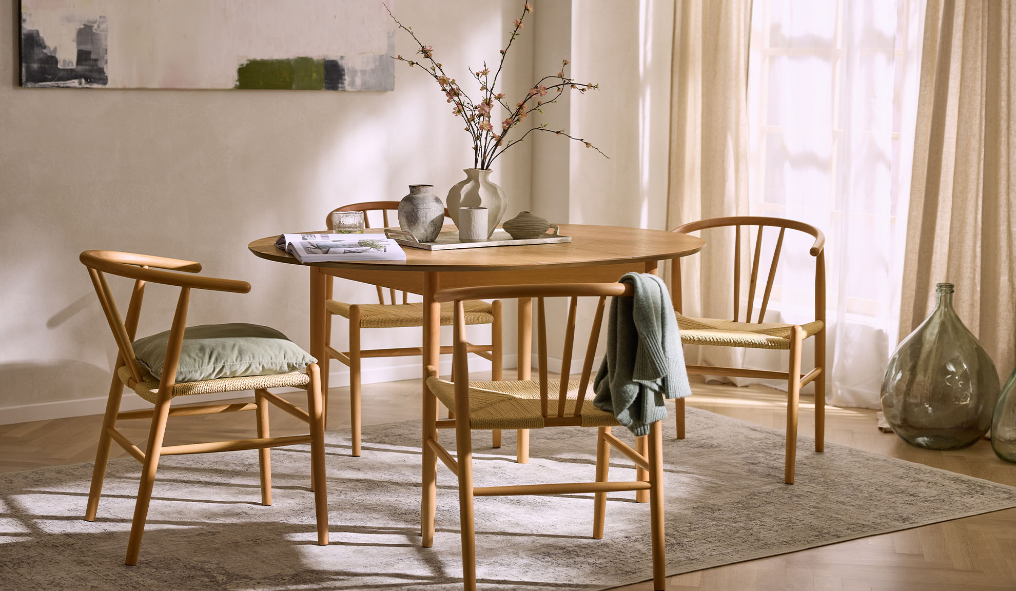 Светла трапезария с кръгла дървена маса, дървени столове с въжени седалки и флорална декорация.