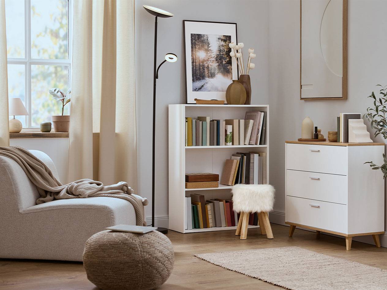 Уютна всекидневна с фотьойл, лавица за книги и скрин в неутрални цветове.