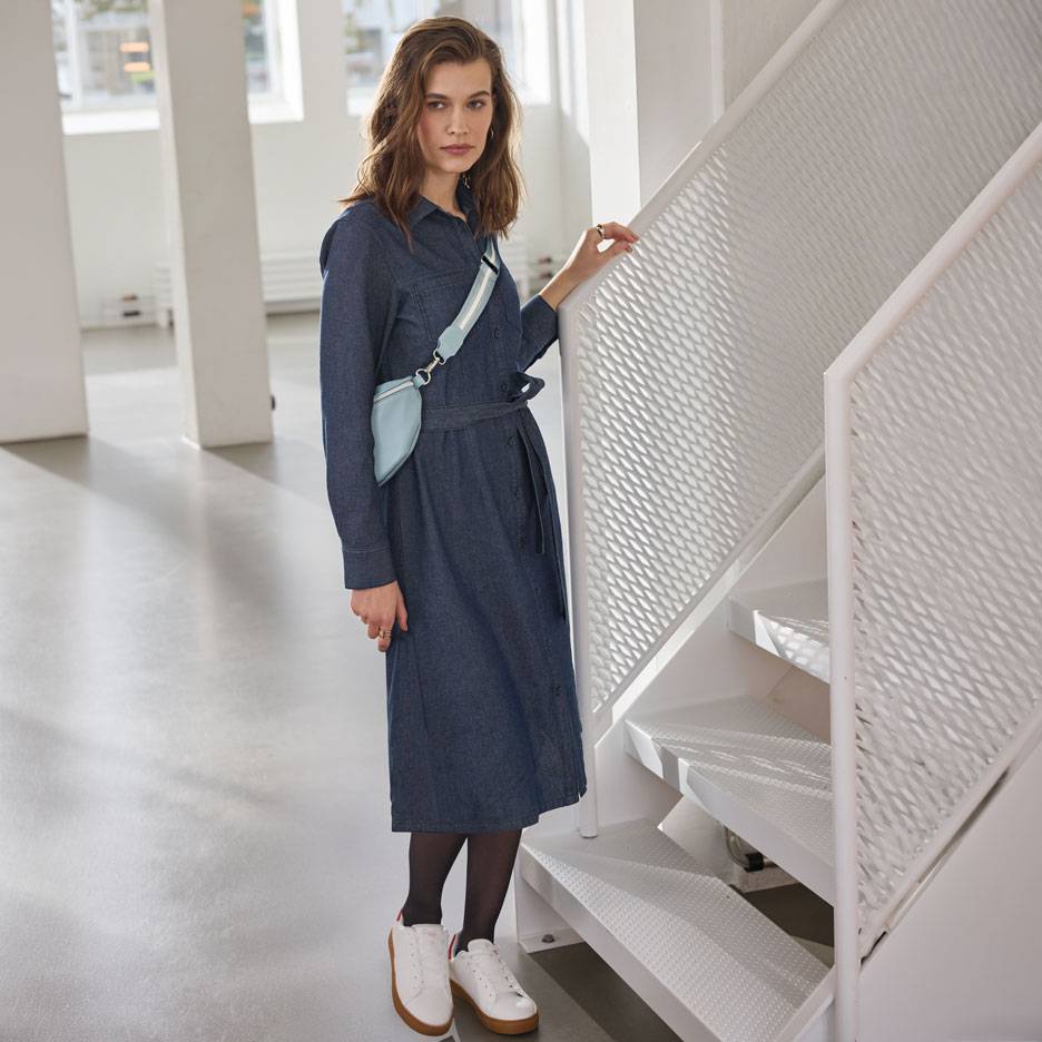 Жена в дънкова рокля-риза, черен чорапогащник и бели маратонки, със светлосиня чанта за кръст.